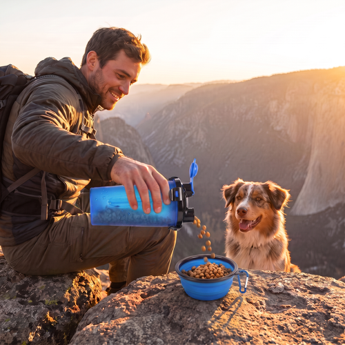 A man hiking outdoors at sunset pours dry dog food from a blue portable container into a blue bowl on a rocky cliffside, with a dog standing nearby and mountain cliffs in the background.